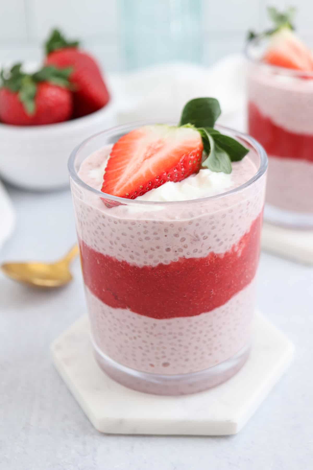 A layered strawberry chia pudding in a glass, topped with a fresh strawberry slice. The pudding has distinct layers of chia mixture and strawberry puree. A bowl of strawberries and a spoon are in the background on a light surface.