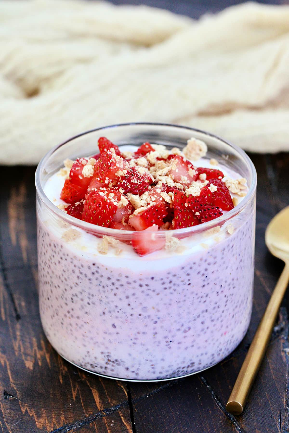 A glass cup filled with chia pudding topped with chopped strawberries and crumbled granola. The dessert is placed on a wooden surface with a gold spoon beside it. A beige textured fabric is in the background.