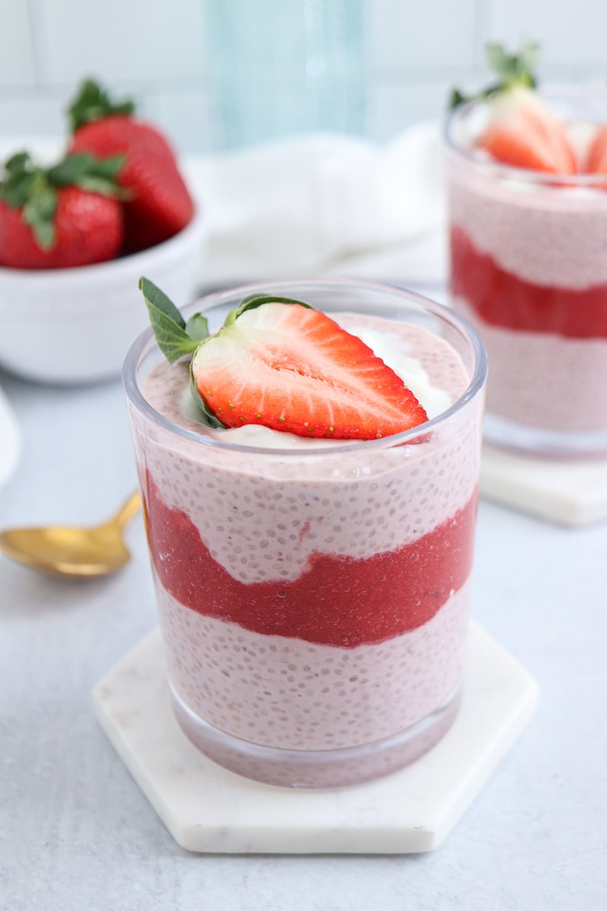 A glass of pink chia pudding layered with a bright red strawberry puree, topped with a halved strawberry. Another glass is partly visible in the background, with fresh strawberries in a small bowl beside it. A gold spoon lies nearby.
