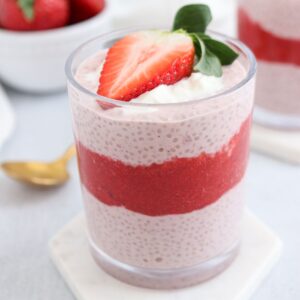 A glass filled with layered strawberry chia pudding. The bottom and top layers are light pink chia pudding, with a middle layer of red strawberry puree. A fresh strawberry slice and a dollop of cream garnish the top. A bowl of strawberries is in the background.