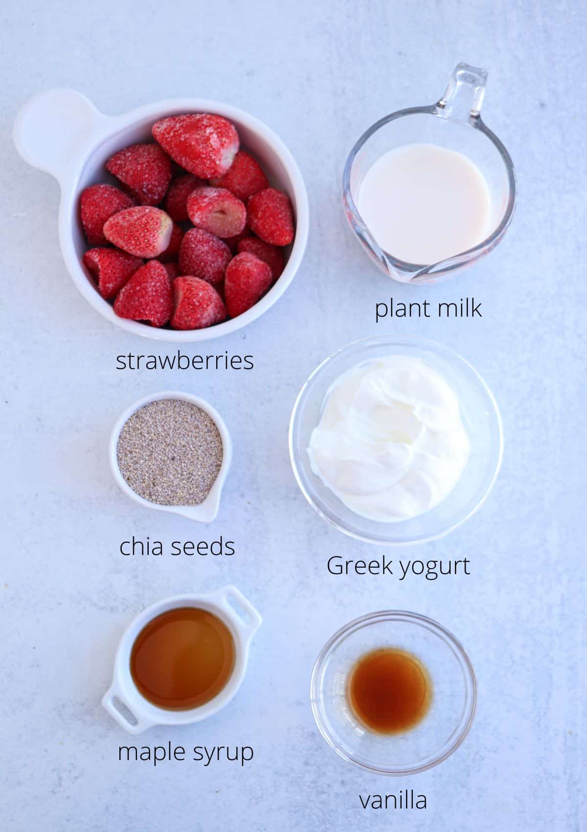 Top view of ingredients: a bowl of frozen strawberries, a cup labeled plant milk, a small bowl of chia seeds, a bowl of Greek yogurt, a small container of maple syrup, and a tiny dish of vanilla, all arranged on a light surface.