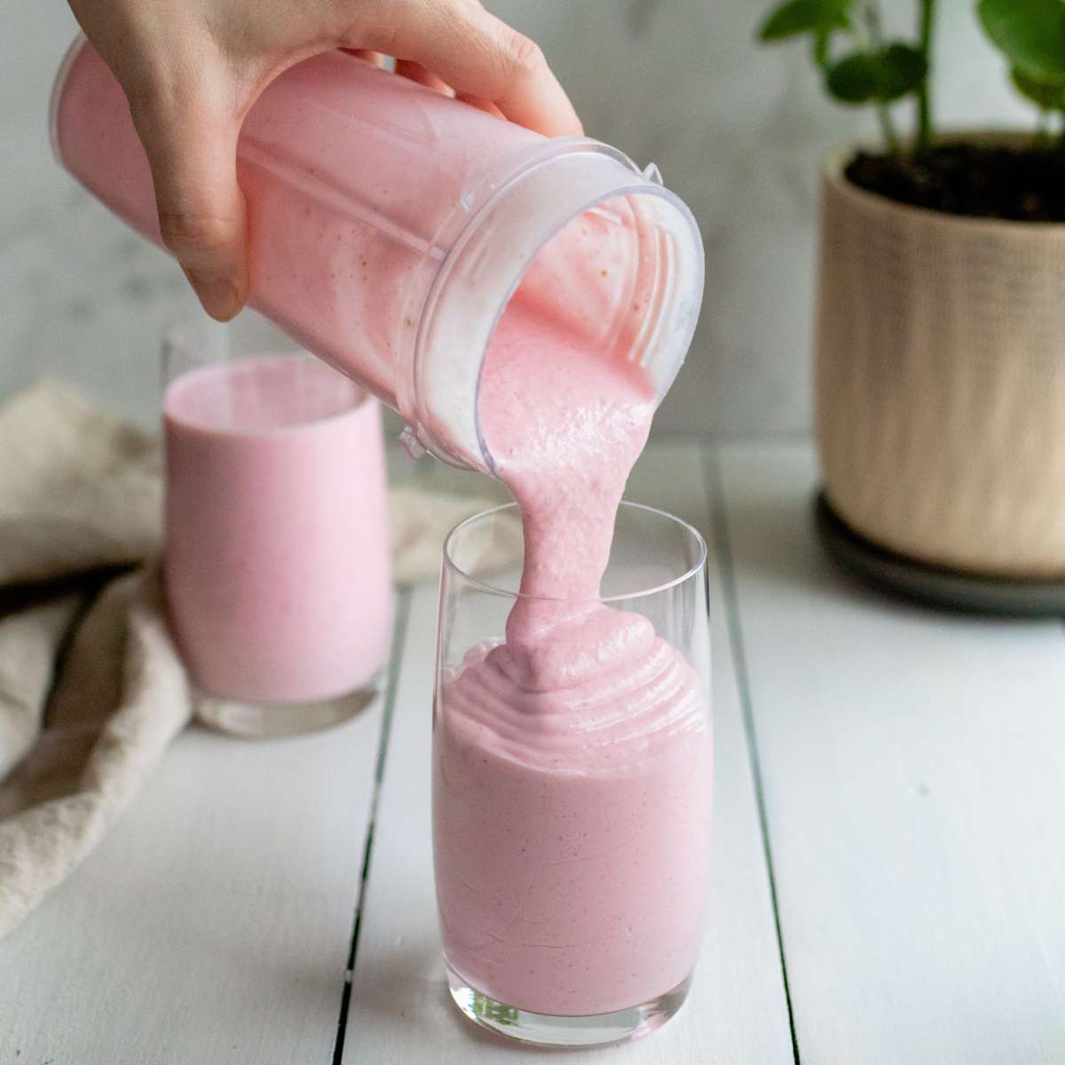 A person pours a thick pink smoothie from a blender cup into a clear glass on a white wooden surface. Another filled glass and a potted plant are in the background, with a beige cloth nearby.