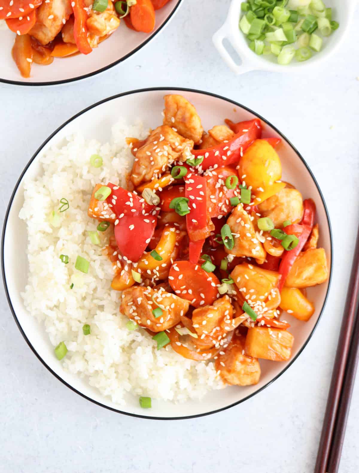 A bowl of sweet and sour chicken served with white rice. The dish includes chicken pieces, red bell peppers, carrots, pineapple chunks, and is garnished with sesame seeds and chopped green onions. A small dish of green onions is on the side.