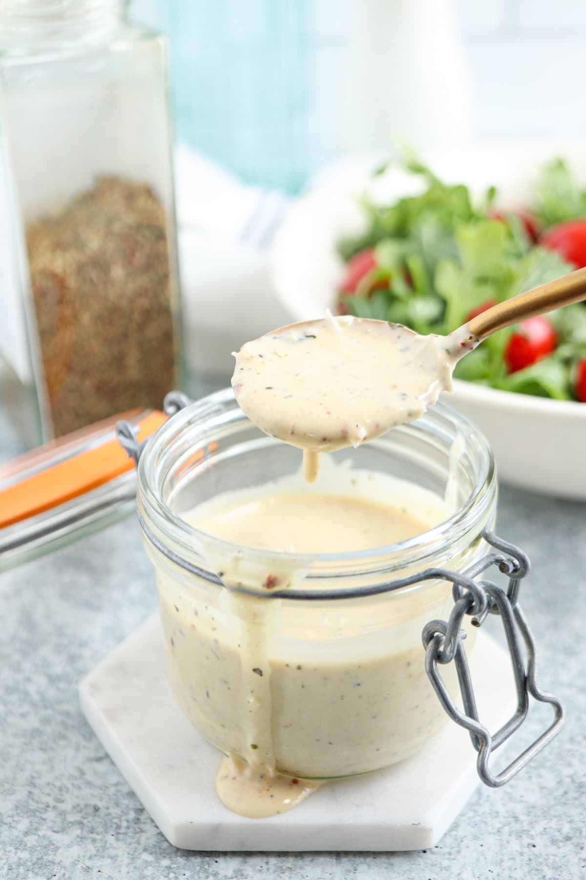 A spoon holds creamy salad dressing above an open glass jar, with dressing dripping back into the jar. A bowl of fresh salad and a jar of herbs are blurred in the background.