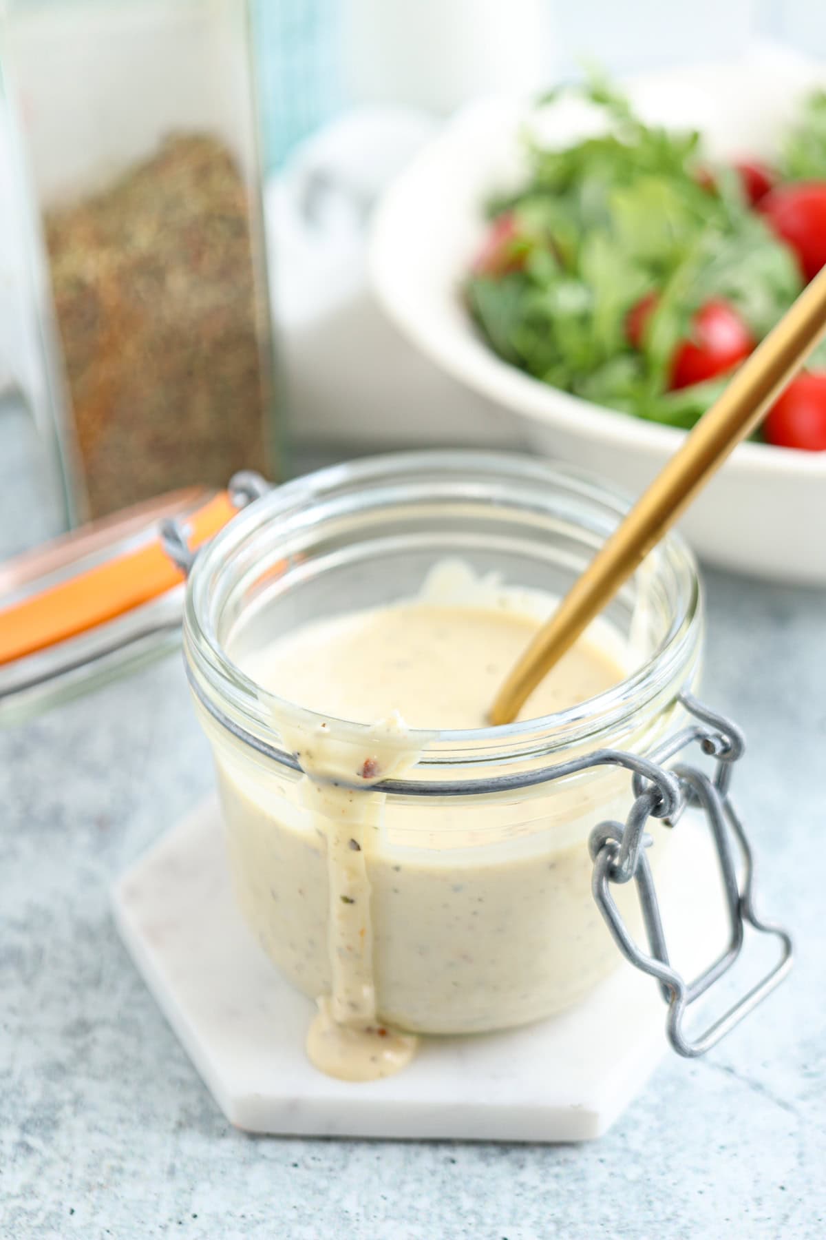 A glass jar of creamy salad dressing with a spoon inside sits on a white hexagonal coaster. Dressing drips down the jar. In the background, there’s a bowl of cherry tomatoes and greens, and a container of spices.