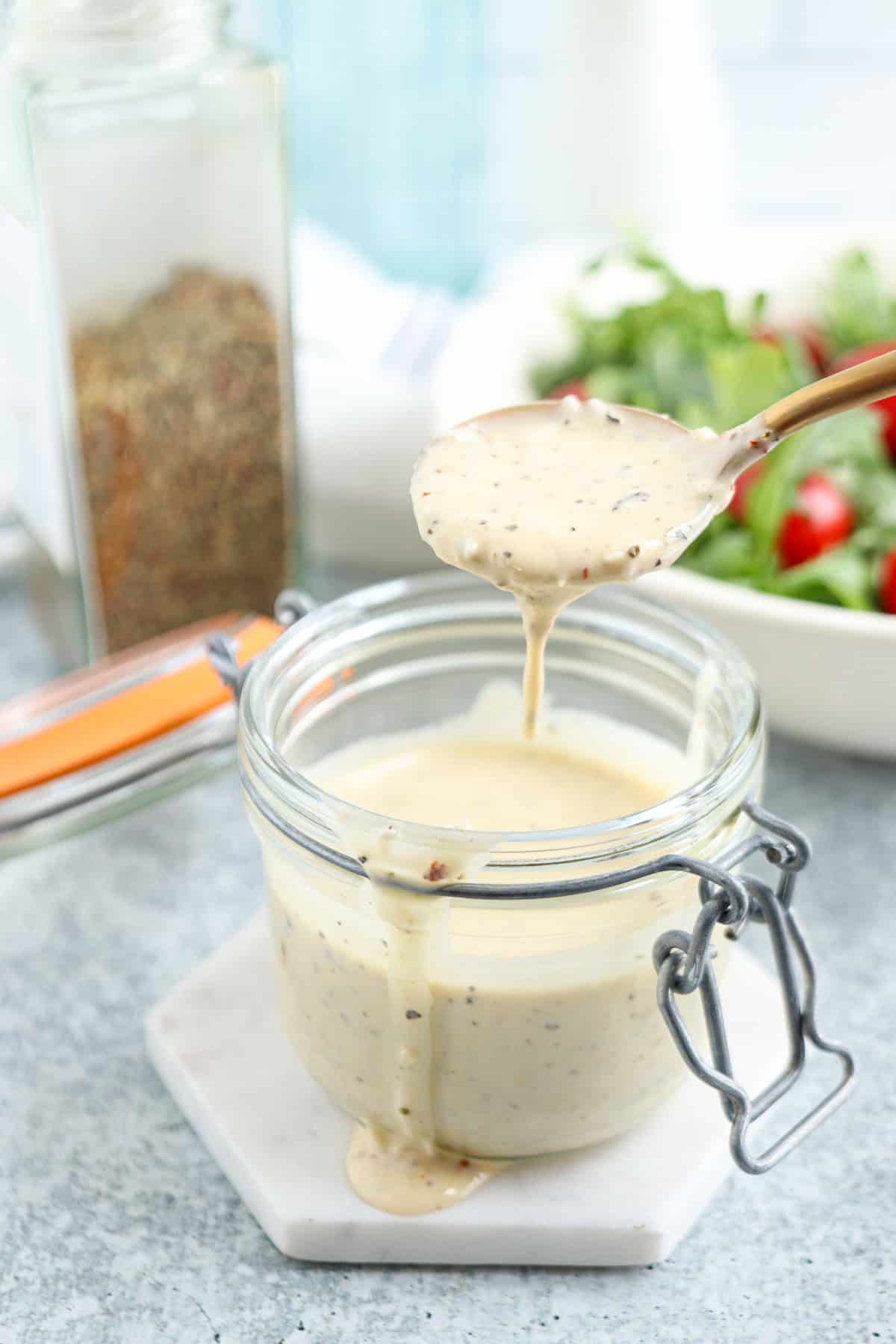 A spoon drizzles creamy, speckled salad dressing into a glass jar on a white coaster, with a salad and spice container blurred in the background.