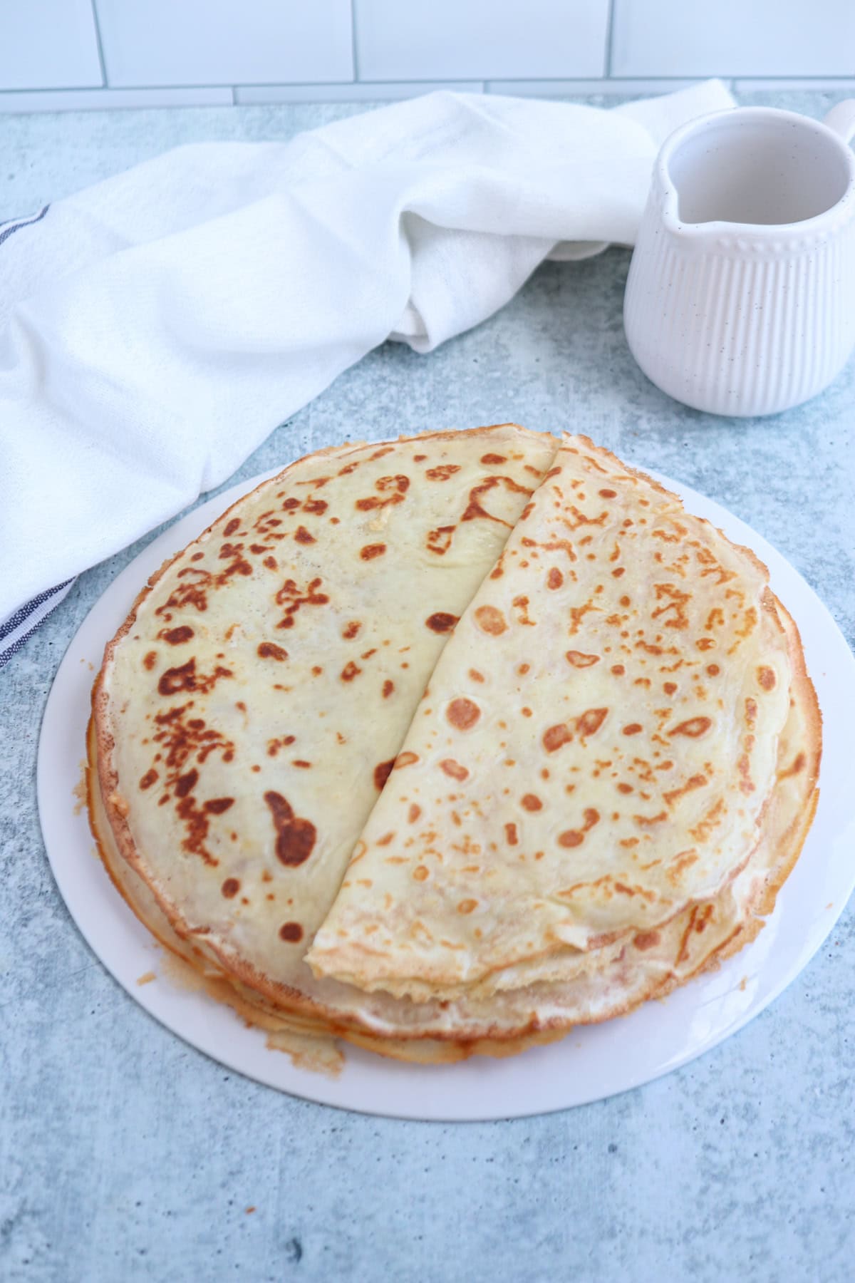 A stack of golden-brown crepes, reminiscent of a classic nalesniki recipe, sits on a white plate, with one crepe artfully folded. A white cloth and a ceramic pitcher adorn the background on a light blue textured surface.