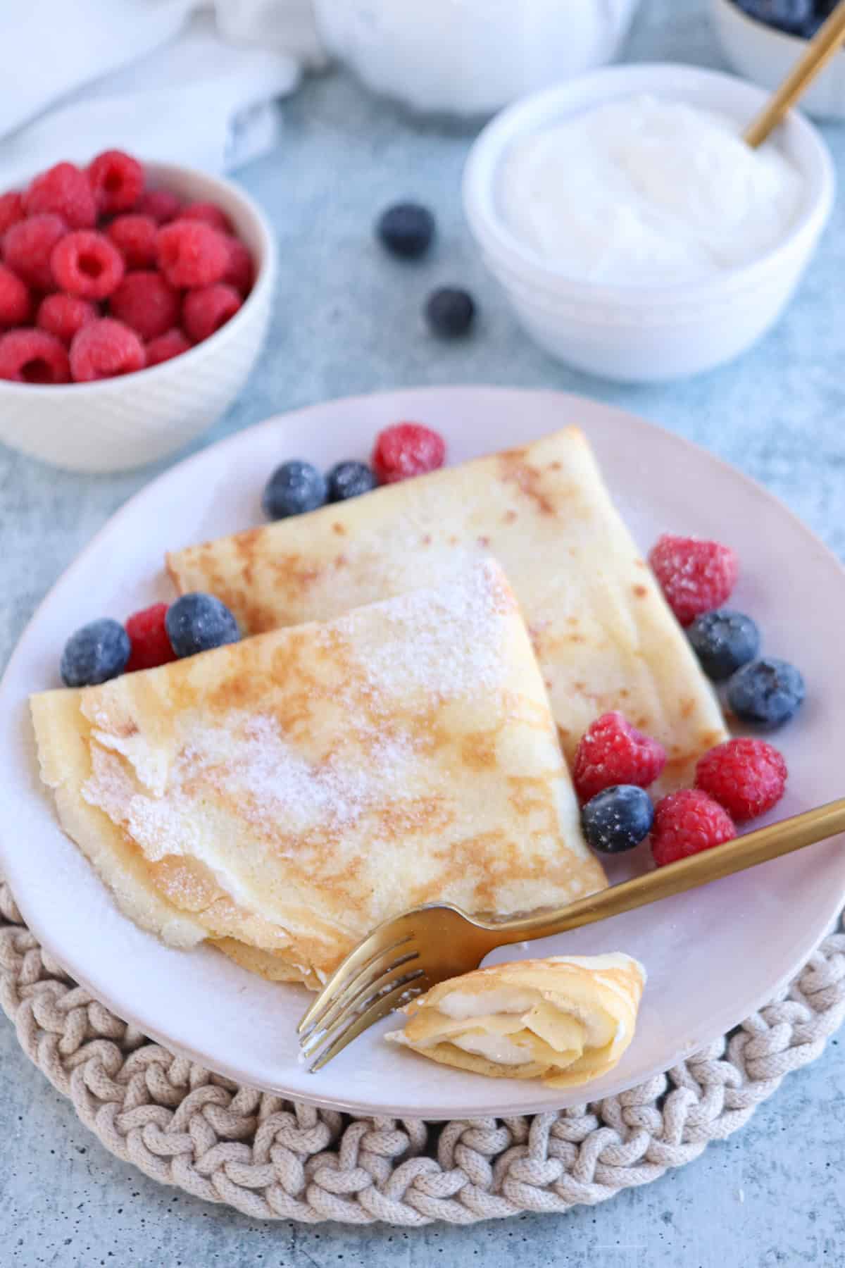 A plate with two folded crepes, reminiscent of a classic nalesniki recipe, is sprinkled with powdered sugar and surrounded by fresh raspberries and blueberries. A fork rests on the plate, while bowls of raspberries and yogurt are visible in the background.