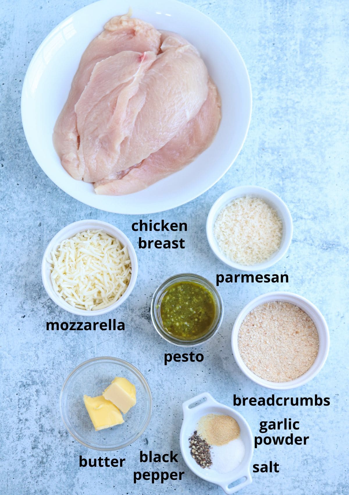 A flat lay of labeled ingredients in bowls for pesto chicken parmesan: raw chicken breast, shredded mozzarella, pesto, grated parmesan, breadcrumbs, garlic powder, salt, black pepper, and two pieces of butter on a light surface.
