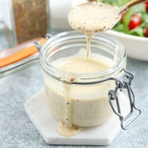 A glass jar filled with creamy salad dressing on a white coaster, with some dressing dripping from a wooden spoon above the jar. A bowl of salad is blurred in the background.