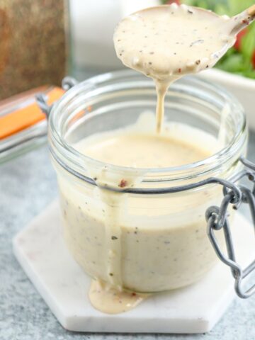 A glass jar filled with creamy salad dressing on a white coaster, with some dressing dripping from a wooden spoon above the jar. A bowl of salad is blurred in the background.