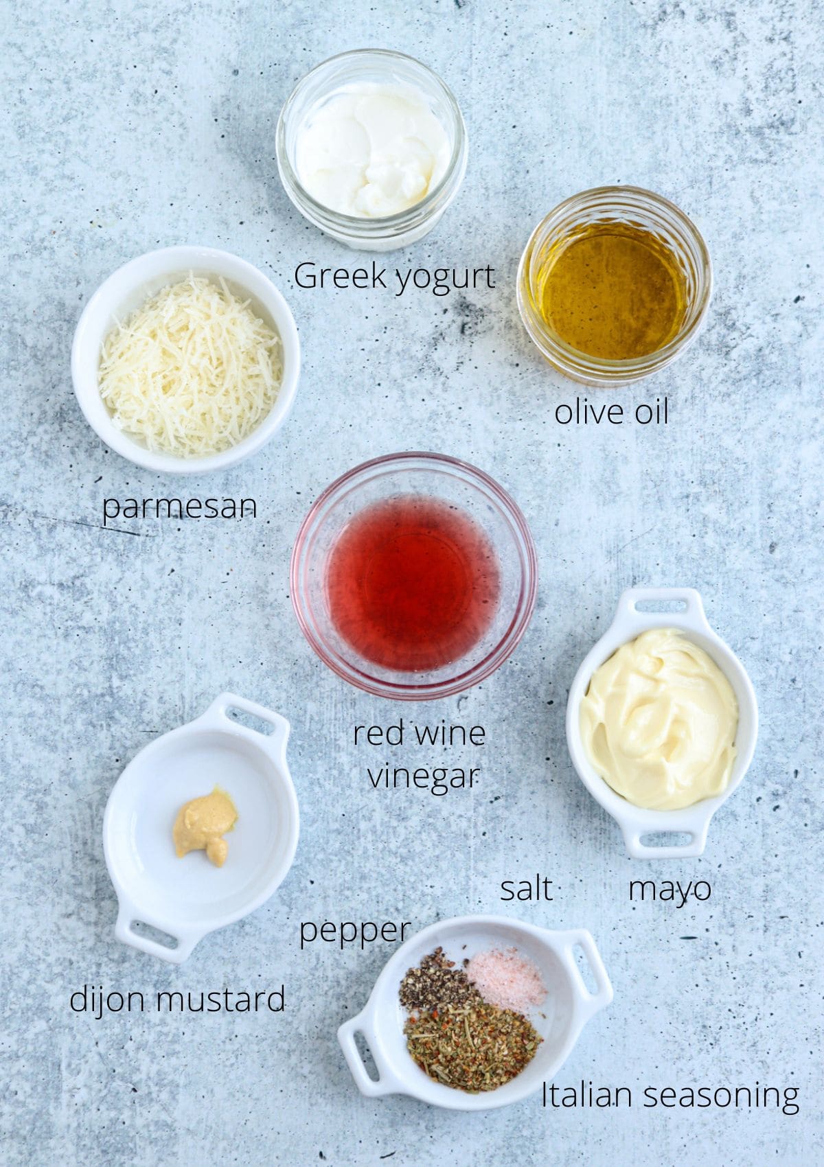 Seven small bowls with labeled ingredients on a gray surface: Greek yogurt, olive oil, parmesan cheese, red wine vinegar, mayo, Dijon mustard, pepper, salt, and Italian seasoning.