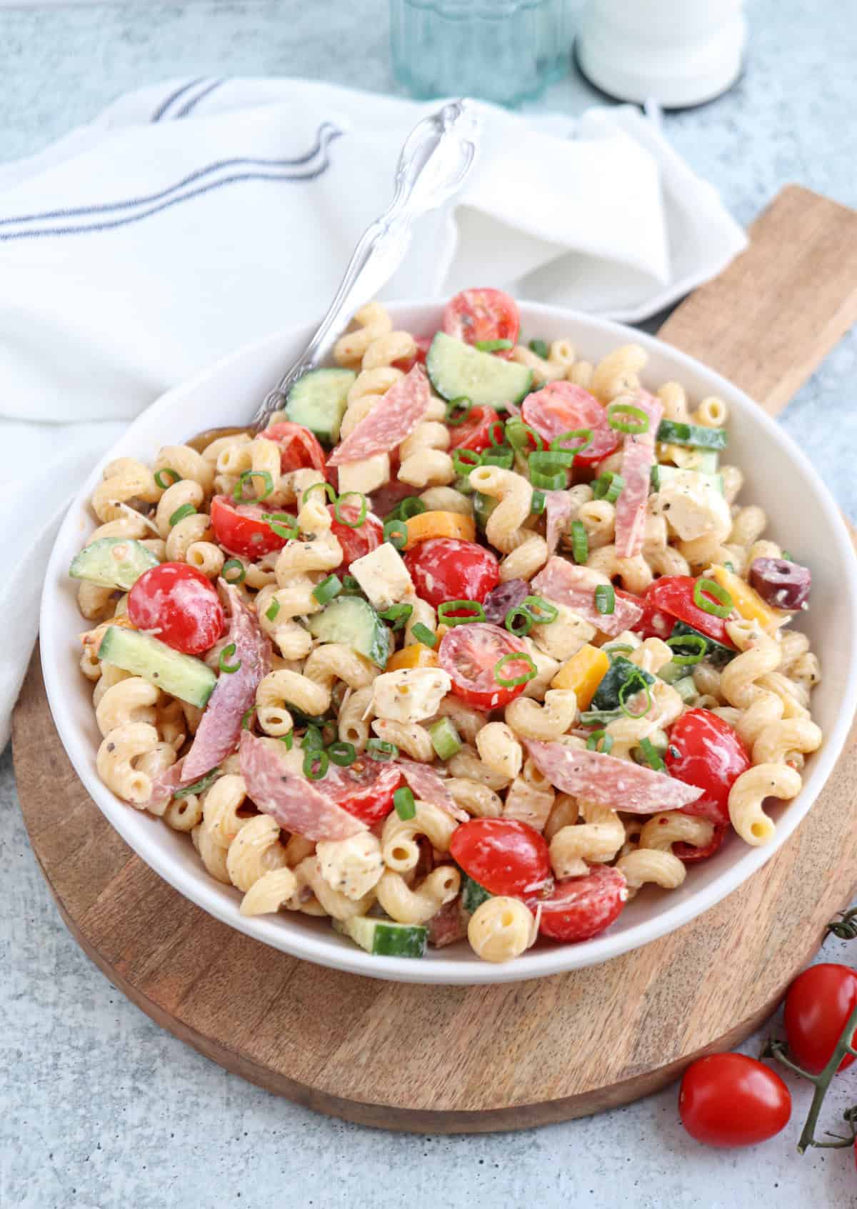 A bowl of pasta salad with curly noodles, cherry tomatoes, cucumber slices, salami strips, green onions, and cubes of cheese on a wooden board, with a fork and a white napkin nearby.