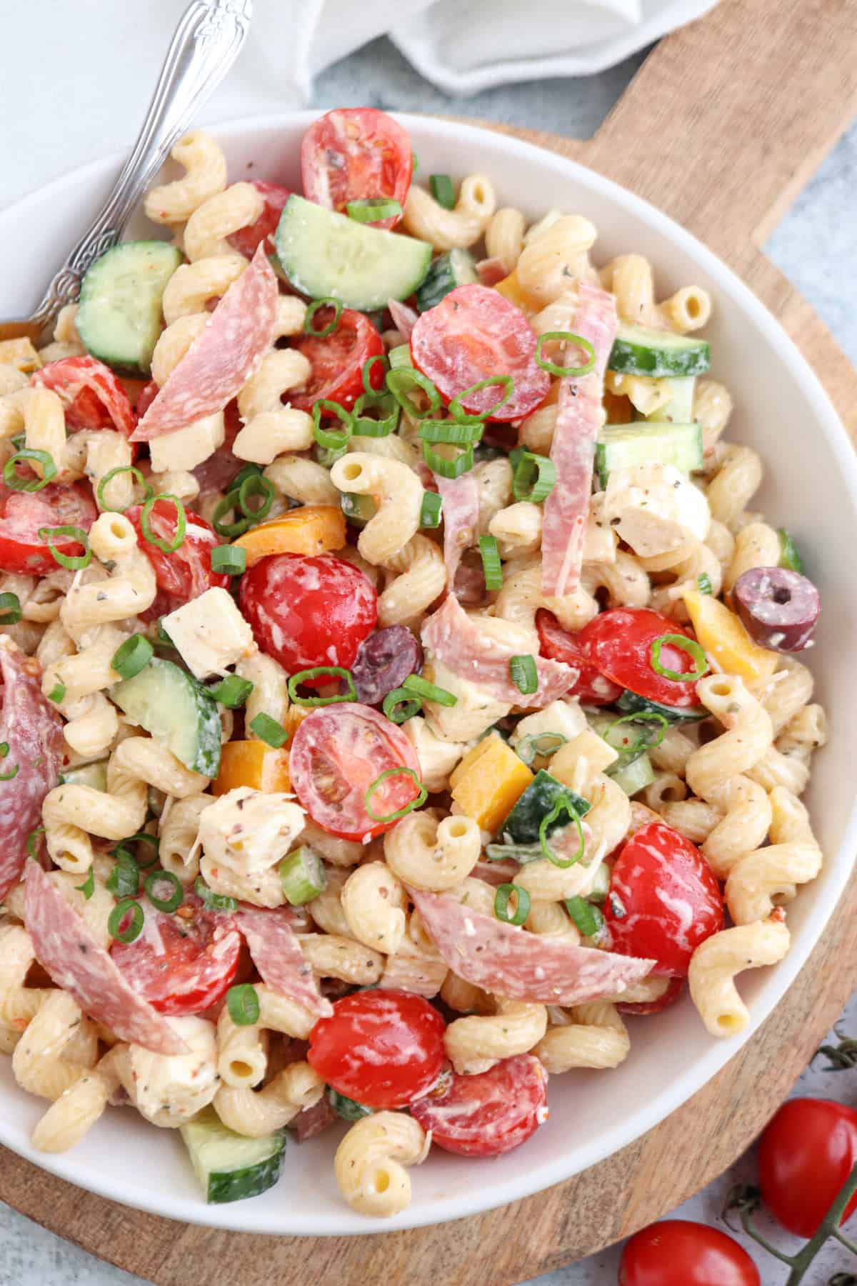 A bowl of pasta salad with curly noodles, cherry tomatoes, cucumber slices, bell peppers, black olives, cubed cheese, salami strips, and chopped green onions, dressed in a creamy sauce.