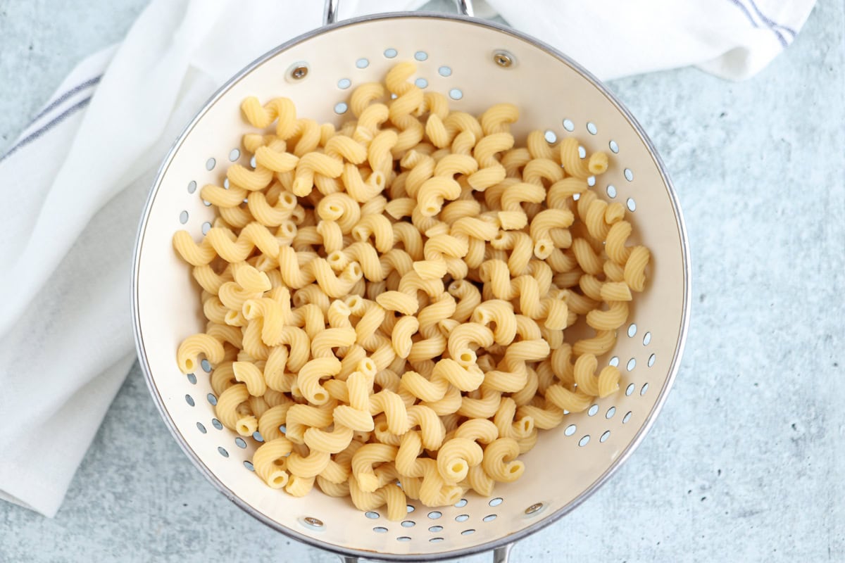A metal colander filled with cooked cavatappi pasta sits on a light blue surface, with a white kitchen towel partially visible in the background.