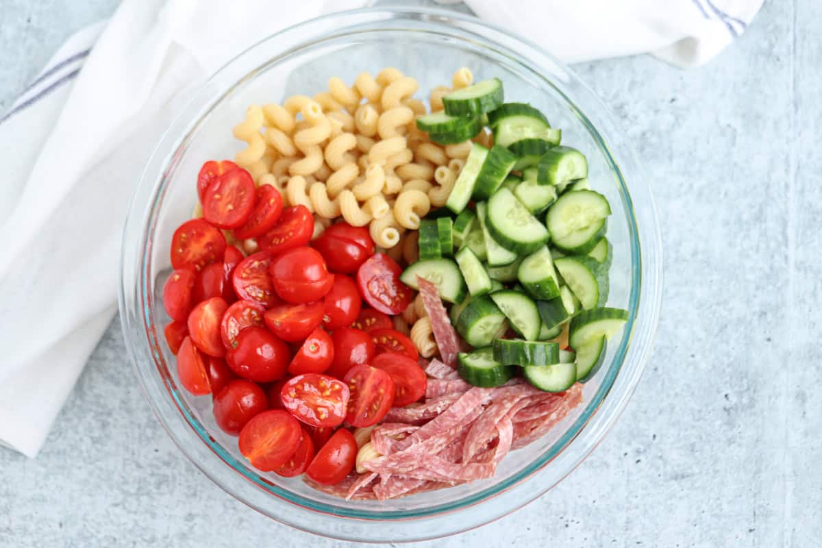 A glass bowl filled with cherry tomatoes, sliced cucumbers, cooked pasta spirals, and thin salami strips, all separated into sections, on a light surface next to a white cloth.
