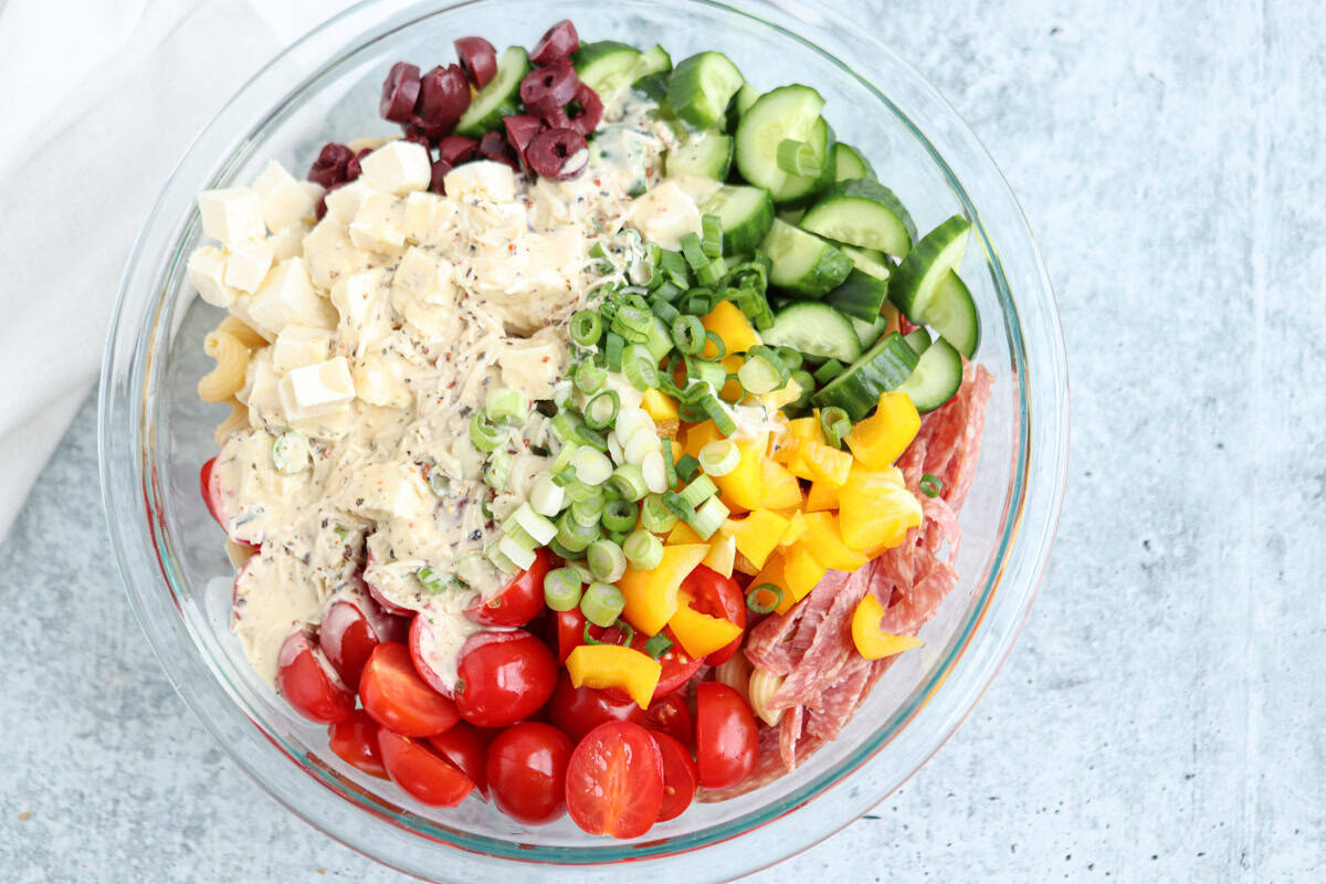 A glass bowl filled with chopped vegetables, sliced cucumbers, cherry tomatoes, diced yellow bell pepper, green onions, kalamata olives, cubed cheese, and salami, topped with a creamy dressing.