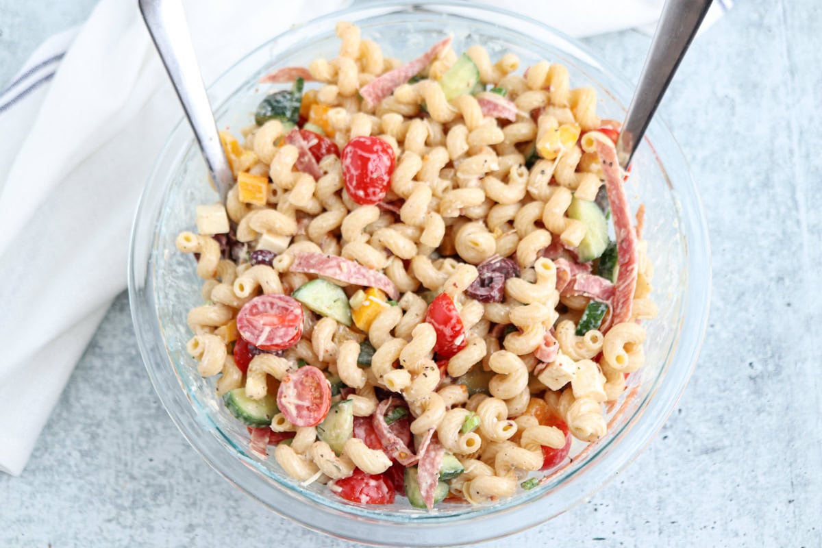 A glass bowl filled with creamy pasta salad, containing curly pasta, cherry tomatoes, cucumber, olives, sliced salami, and cheese, with two serving utensils.