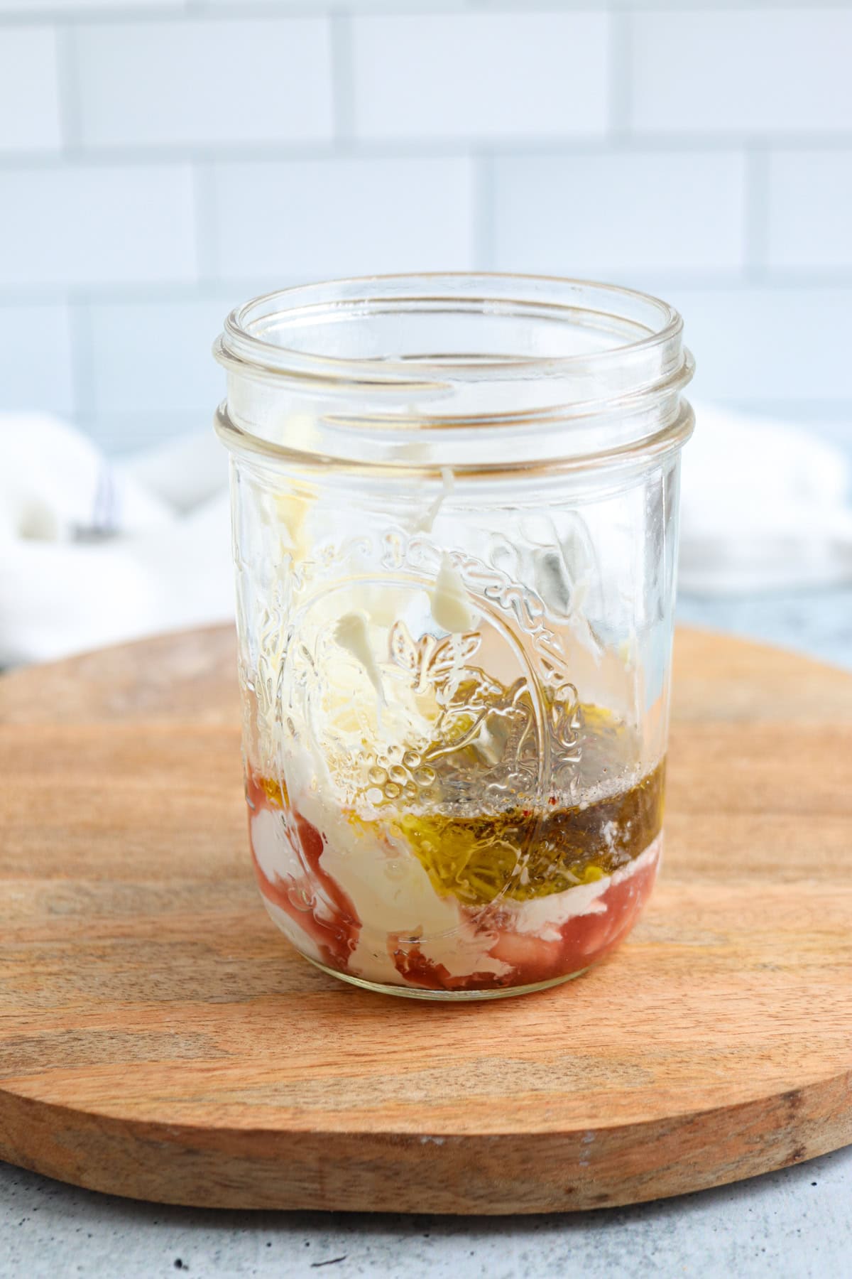A glass jar on a wooden board contains layers of mayonnaise, ketchup, and oil, with a white tiled background.