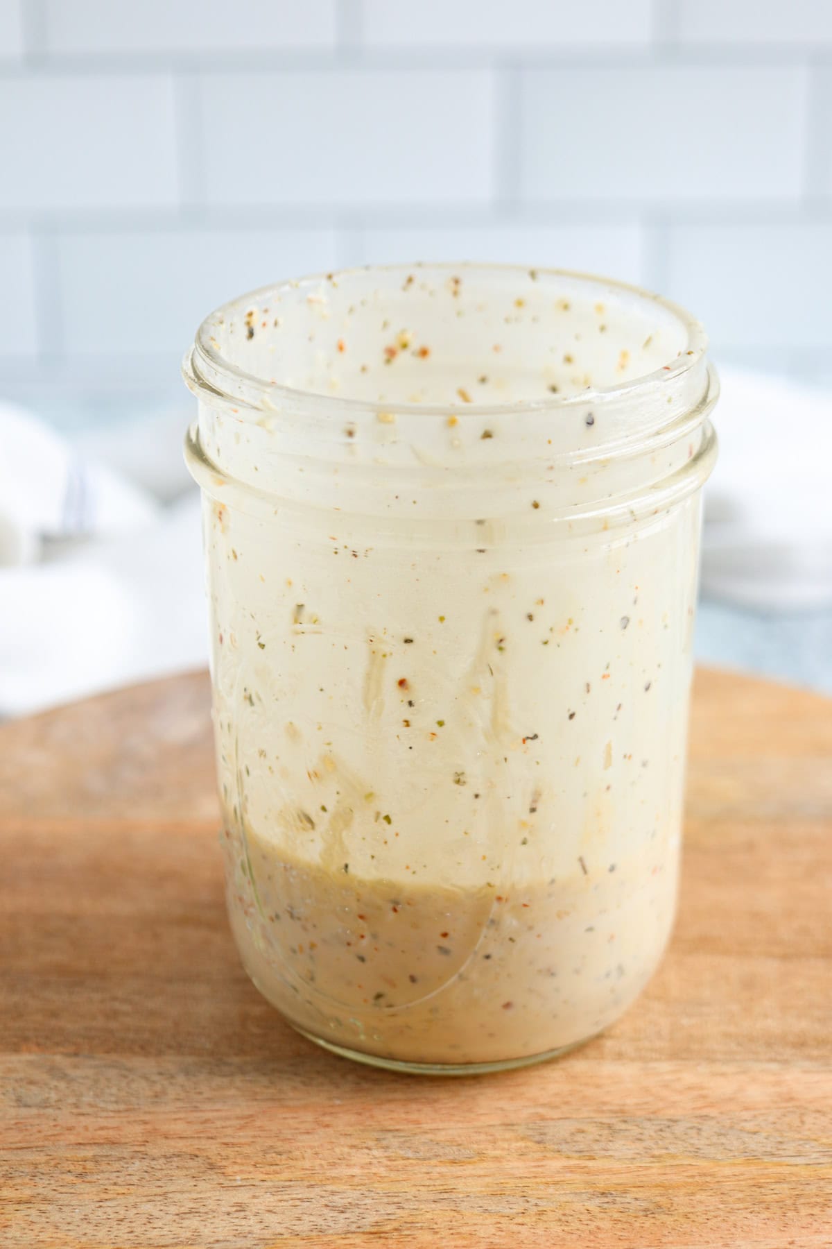 A glass jar with creamy salad dressing, herbs, and spices visible, sits on a wooden surface against a tiled white background.