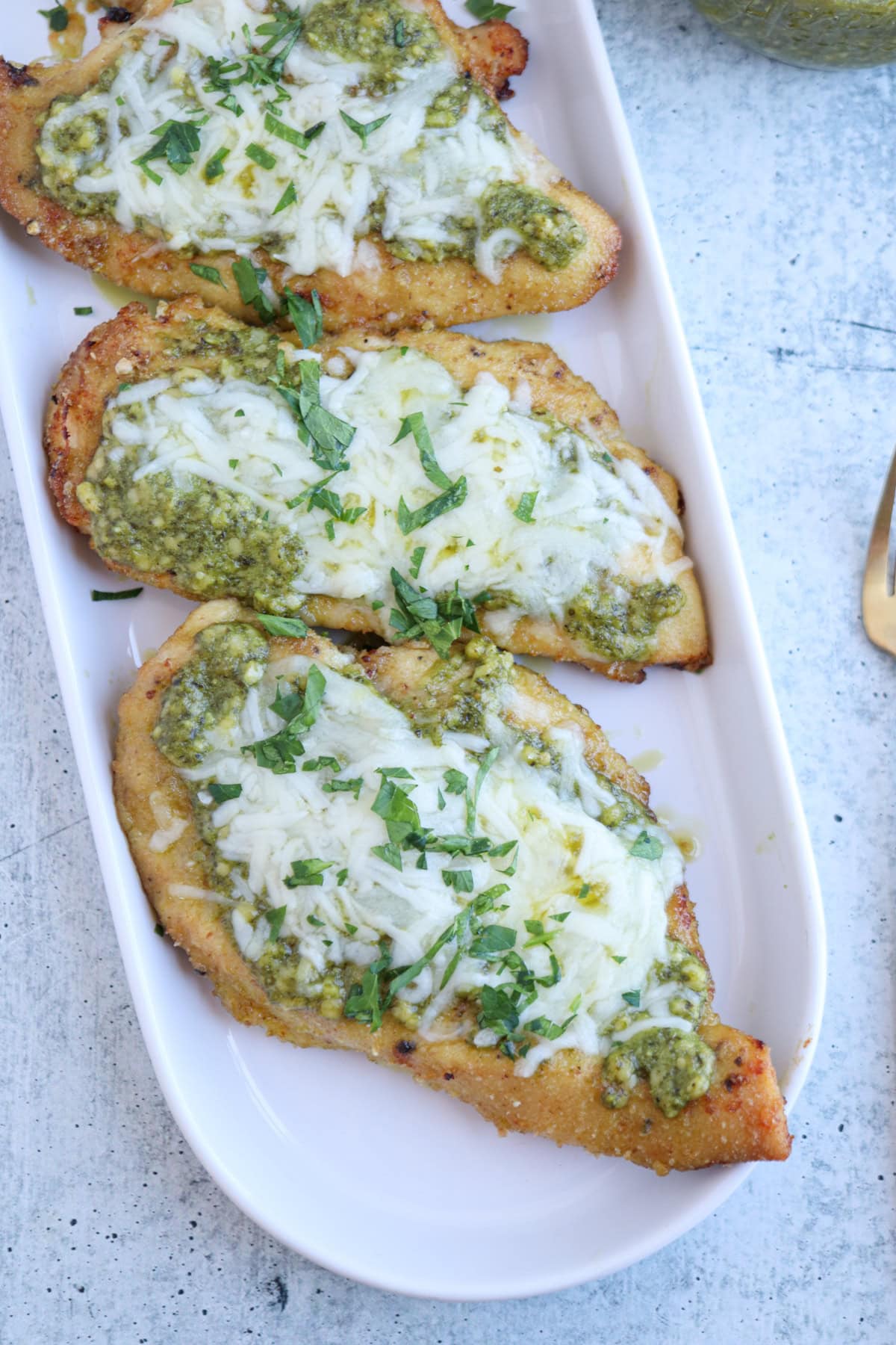Three pieces of cooked pesto chicken parmesan breast topped with melted cheese, green pesto, and chopped fresh herbs are arranged on a white oval plate.