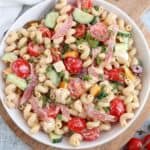 A bowl of pasta salad with curly noodles, cherry tomatoes, cucumber, olives, sliced salami, yellow bell pepper, and green onions, all tossed in a creamy dressing.