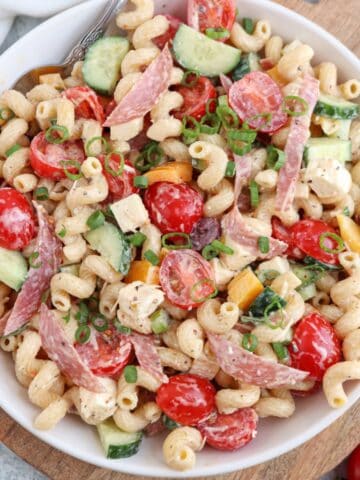 A bowl of pasta salad with curly noodles, cherry tomatoes, cucumber, olives, sliced salami, yellow bell pepper, and green onions, all tossed in a creamy dressing.