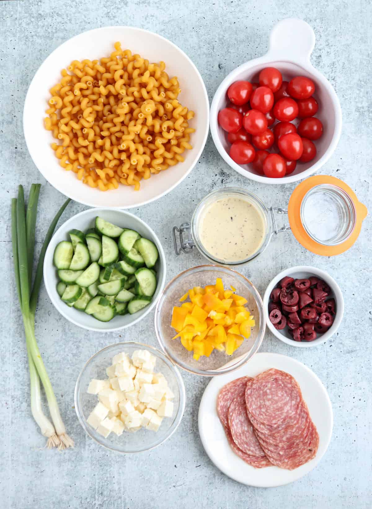 A top-down view of pasta, cherry tomatoes, sliced cucumber, yellow bell pepper, kalamata olives, feta cheese, green onions, salad dressing, water, and sliced salami arranged in separate bowls on a light surface.
