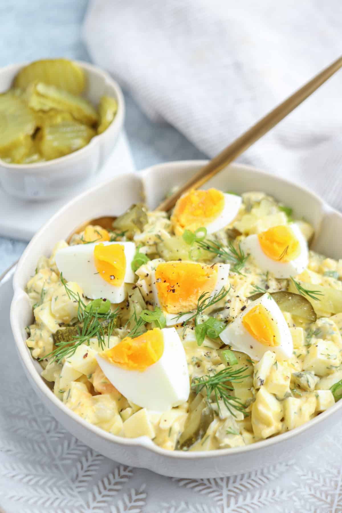 A bowl of creamy egg salad garnished with quartered boiled eggs, fresh dill, and green onions, with a gold spoon. A small dish of sliced pickles is in the background, set on a light surface with a white napkin.