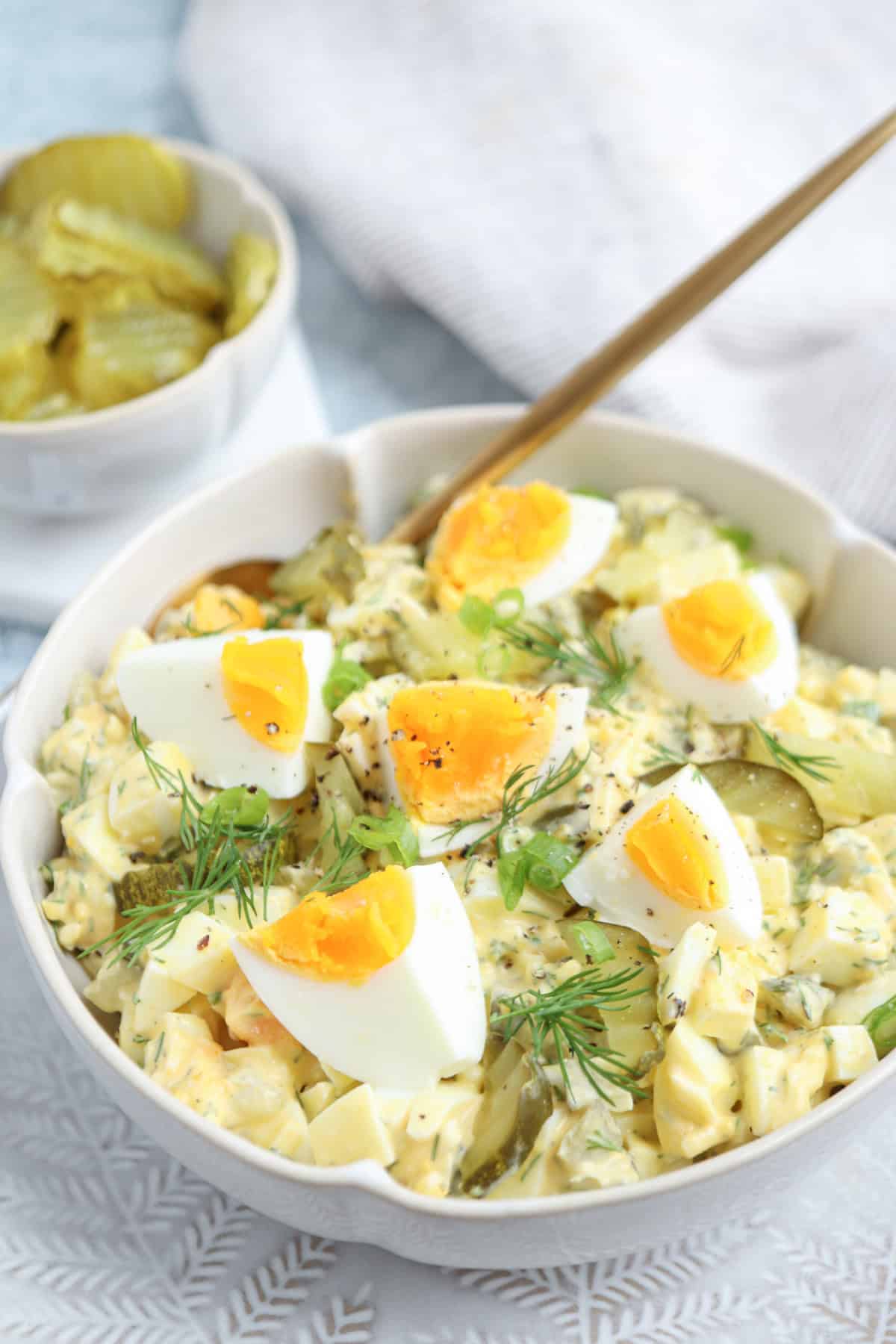A bowl of creamy potato salad topped with quartered boiled eggs, fresh dill, and green onions, with a fork resting in the bowl. A small dish of sliced pickles is in the background.