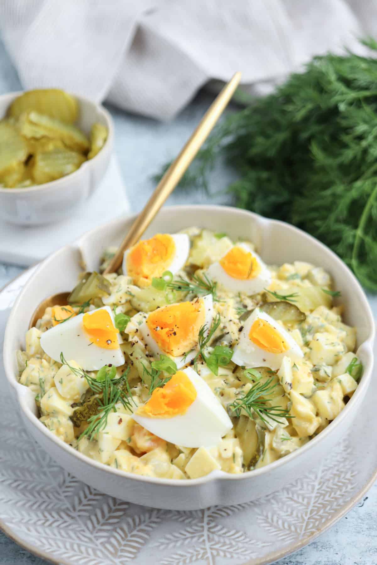 A bowl of creamy egg salad garnished with fresh dill and quartered boiled eggs, with slices of pickles on top. A fork rests in the bowl, and fresh dill and a small dish of pickles are nearby.