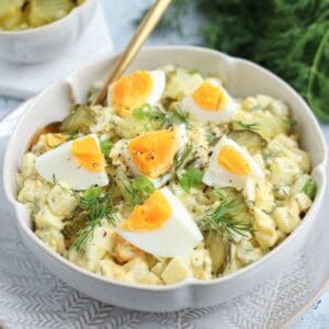 A bowl of creamy egg salad topped with quartered boiled eggs, fresh dill, black pepper, and sliced green onions, served with a gold spoon on a patterned plate. Pickles are visible in the background.