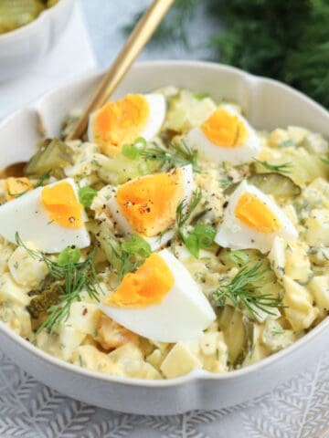 A bowl of creamy egg salad topped with quartered boiled eggs, fresh dill, black pepper, and sliced green onions, served with a gold spoon on a patterned plate. Pickles are visible in the background.