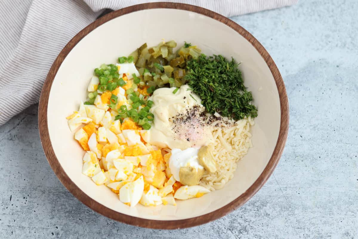 A bowl with chopped hard-boiled eggs, green onions, pickles, fresh dill, shredded cheese, mayonnaise, mustard, and seasonings, ready to be mixed for an egg salad.
