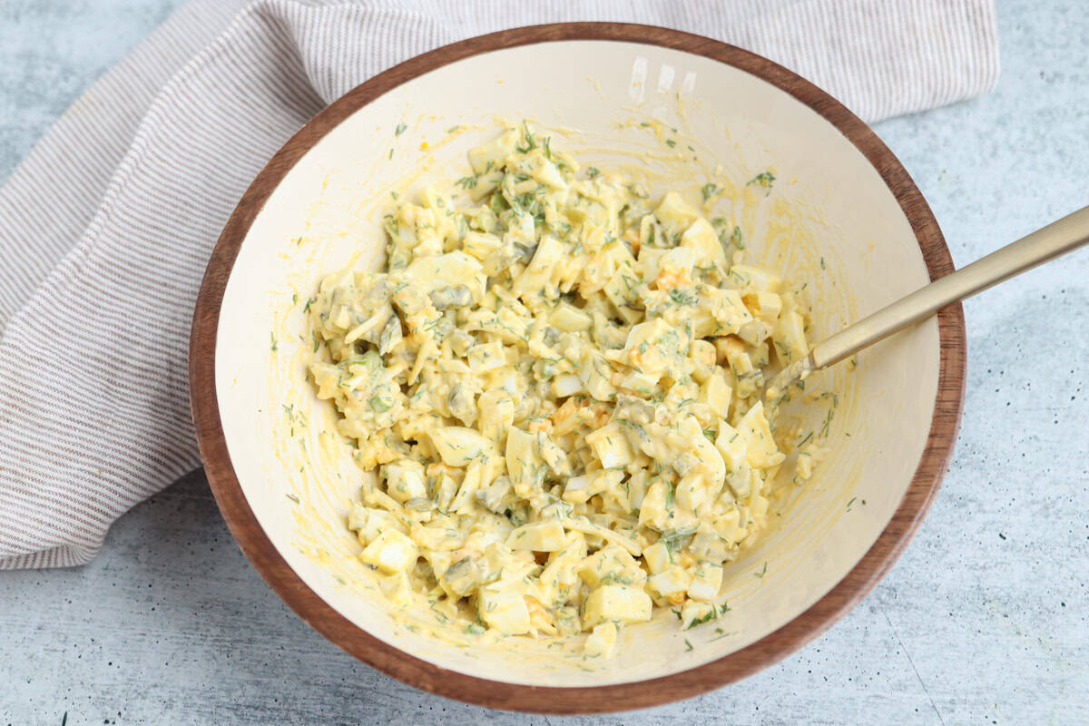 A bowl filled with creamy egg salad, featuring chopped eggs, herbs, and a yellow mayonnaise-based dressing, with a spoon in the bowl. A striped towel lies beside the bowl on a light surface.