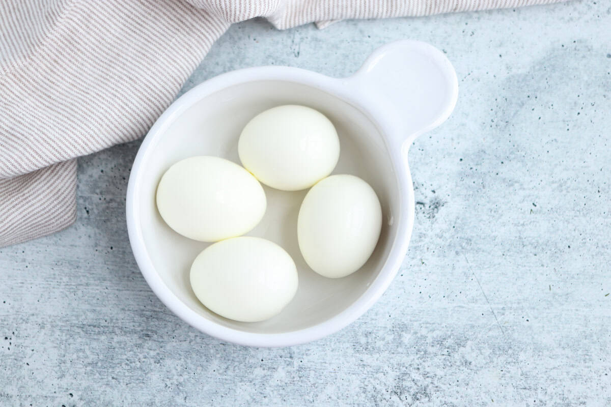 A white bowl containing four hard-boiled eggs sits on a light gray textured surface next to a beige striped cloth.