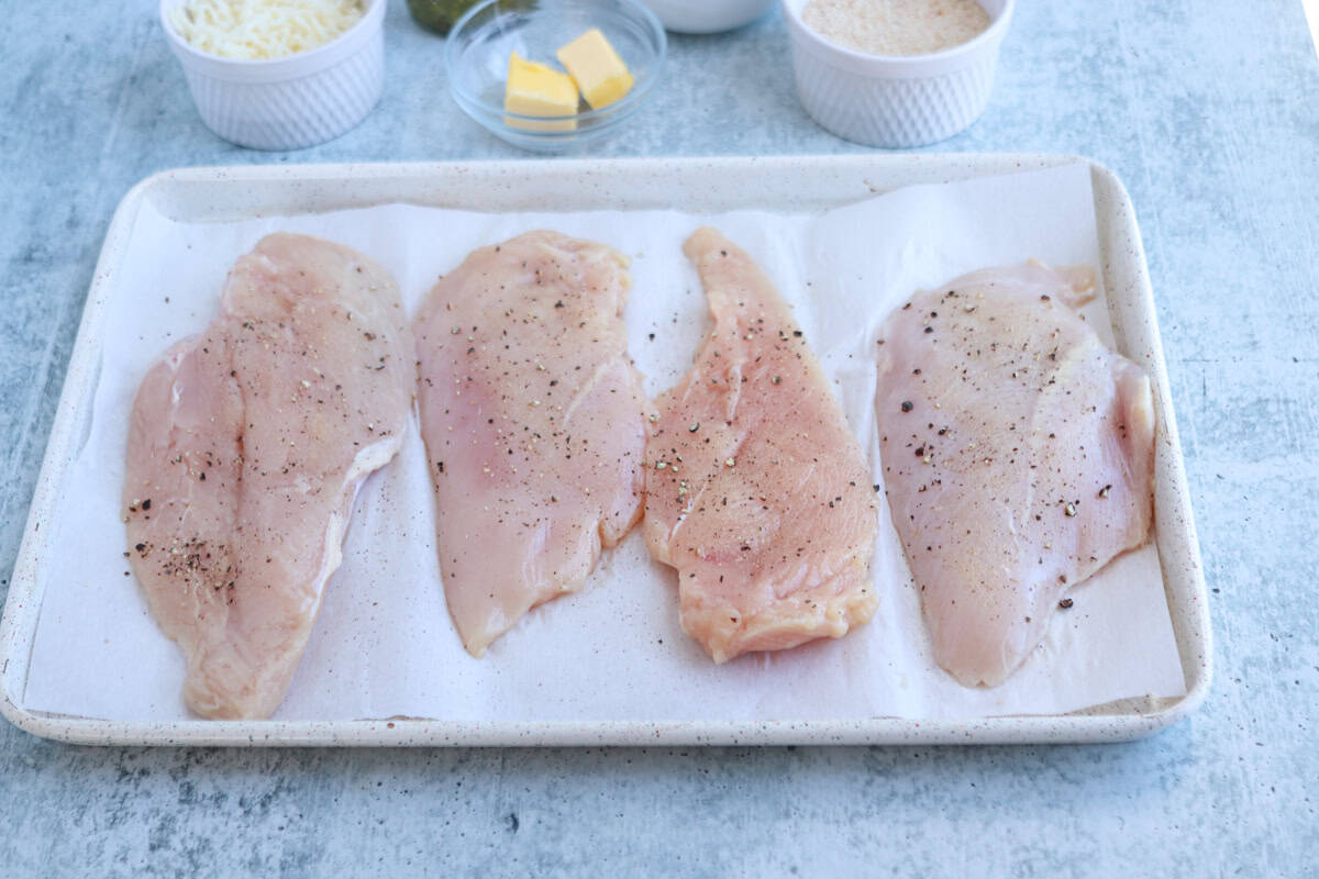 Four raw, seasoned chicken breasts are arranged on a parchment-lined tray for pesto chicken parmesan, with small bowls of shredded cheese, butter, and breadcrumbs visible in the background on a light blue surface.