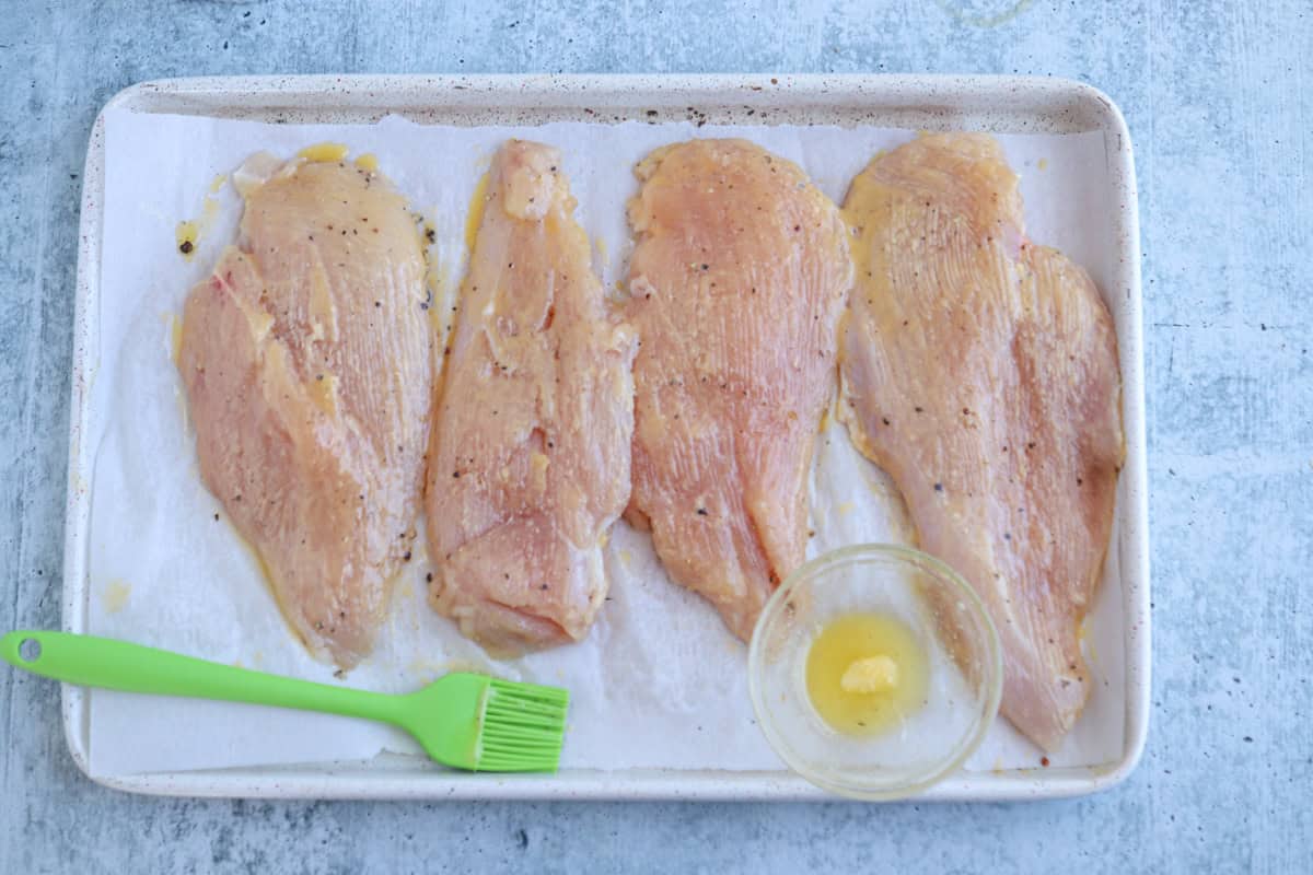 Four raw, seasoned chicken breasts are laid out on a parchment-lined baking tray, ready for the oven. A green brush and a small bowl of melted butter hint at prepping a delicious pesto chicken parmesan dish.