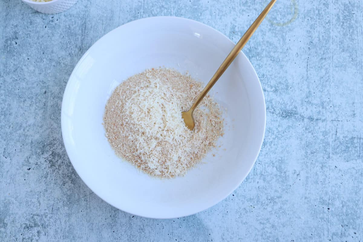 A white bowl containing a mixture of breadcrumbs and grated cheese for pesto chicken parmesan, with a gold-colored fork, set on a light blue textured surface.