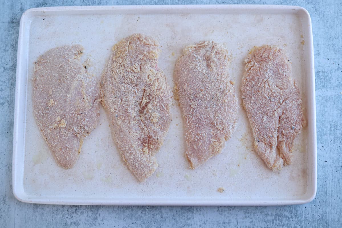 Four breaded raw chicken breasts, perfect for making pesto chicken parmesan, are evenly spaced on a rectangular white tray, ready for cooking. The tray sits on a light gray textured surface.