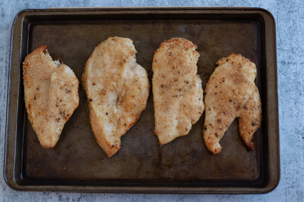 Four breaded and baked pesto chicken parmesan cutlets are arranged in a row on a dark metal baking sheet, set on a light grey textured surface.
