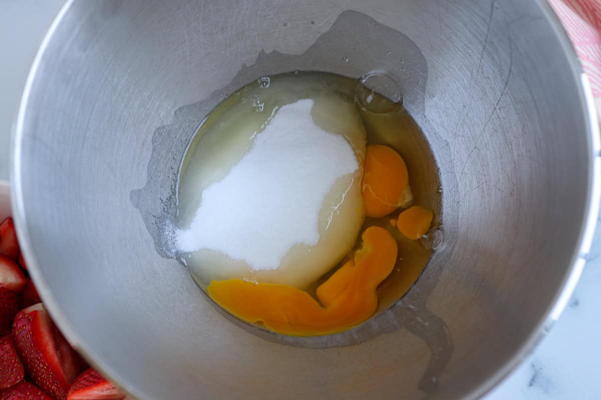 A metal mixing bowl containing three cracked eggs and a pile of white sugar, ready to be mixed for a dessert to pair with pesto chicken parmesan. Sliced strawberries are visible in the corner of the image.
