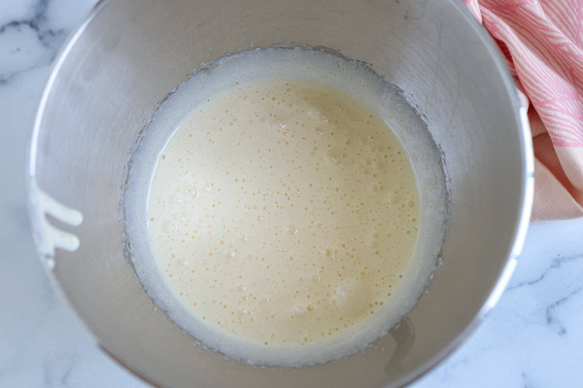A stainless steel mixing bowl containing a pale, frothy batter for pesto chicken parmesan sits on a white marble countertop next to a pink and white striped cloth.