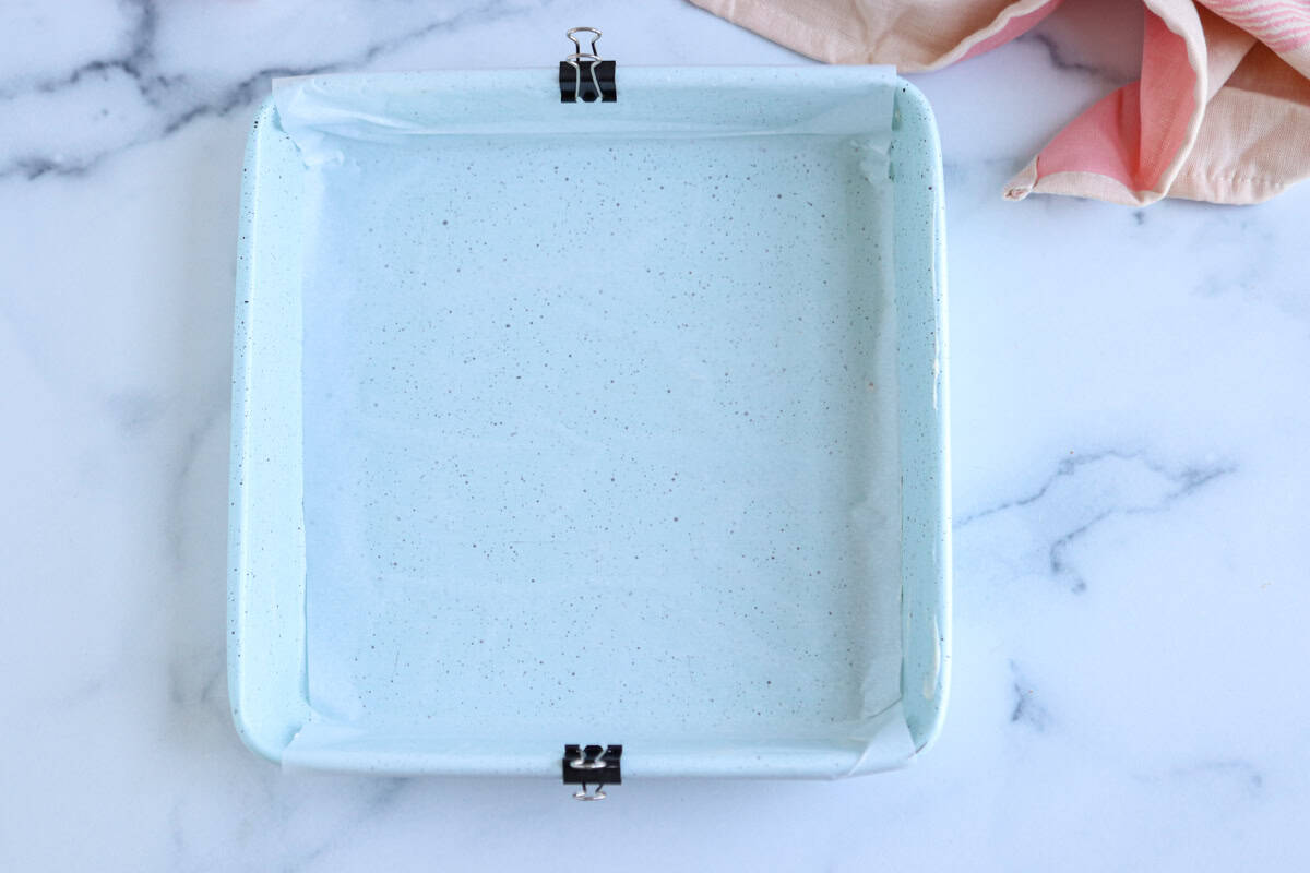 A light blue, square baking dish lined with parchment paper and secured with binder clips at two sides is set on a marble surface, ready for baking pesto chicken parmesan. A pink and beige striped cloth lies in the corner.