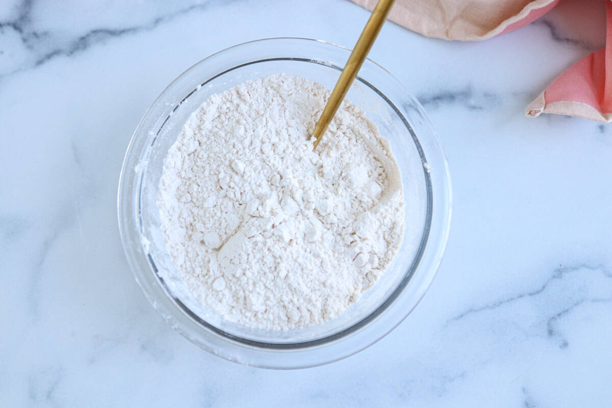 A glass bowl filled with flour sits on a white marble surface, ready for coating pesto chicken parmesan. A gold spoon is partially submerged in the flour, and a pink and beige cloth is visible in the top right corner.