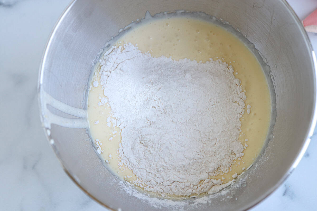 A metal mixing bowl contains a pale yellow batter with a mound of white flour on top, ready to be mixed together for a delicious pesto chicken parmesan dish.