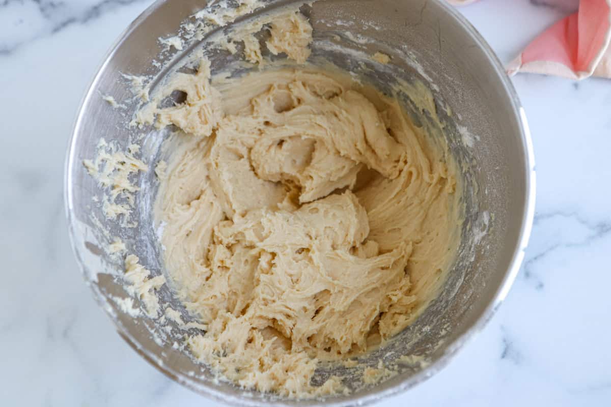 A metal mixing bowl filled with creamy, light-colored pesto chicken parmesan cookie dough sits on a white marble surface. Some dough clings to the sides of the bowl, and a cloth is partially visible in the corner.
