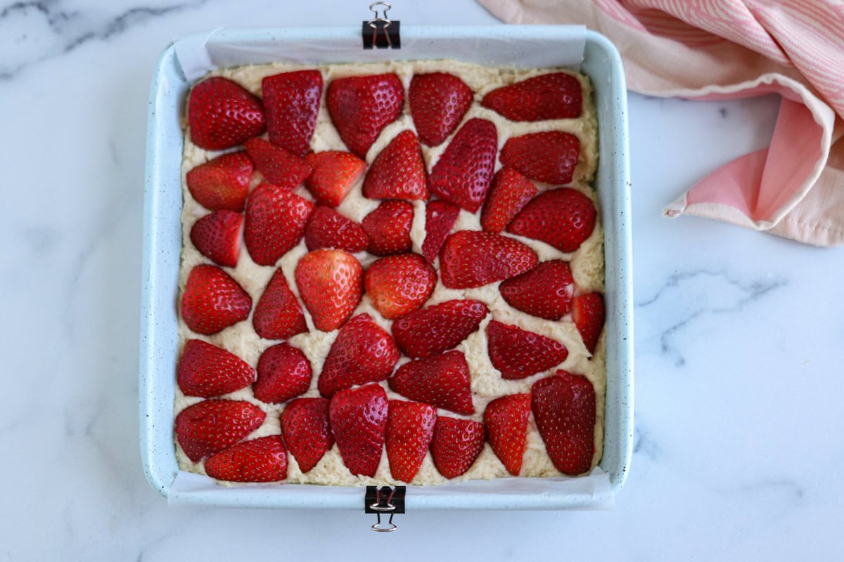 A square baking pan filled with unbaked cake batter, topped with halved fresh strawberries, sits on a marble surface next to a pink and white striped cloth—ready for the oven like a classic pesto chicken parmesan.