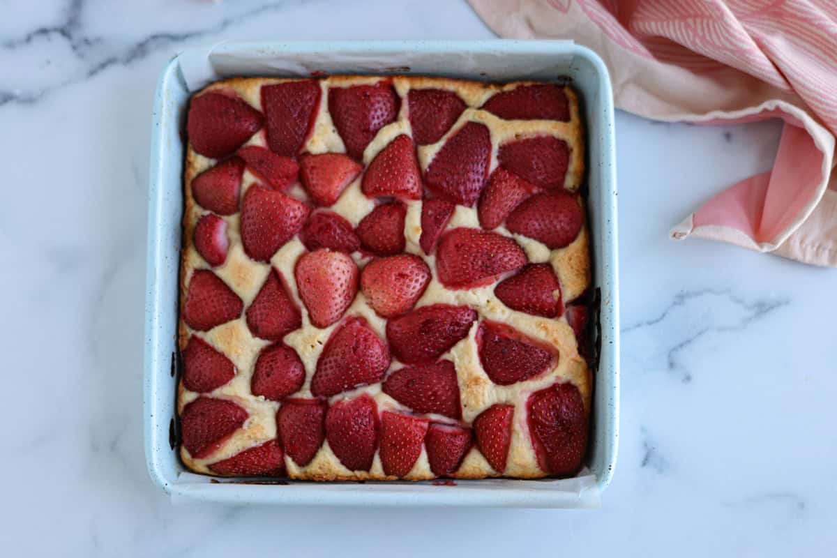 A square baking pan filled with golden cake, reminiscent of the warmth of a pesto chicken parmesan bake, topped with halved strawberries, arranged neatly on a white marble surface with a pink cloth nearby.
