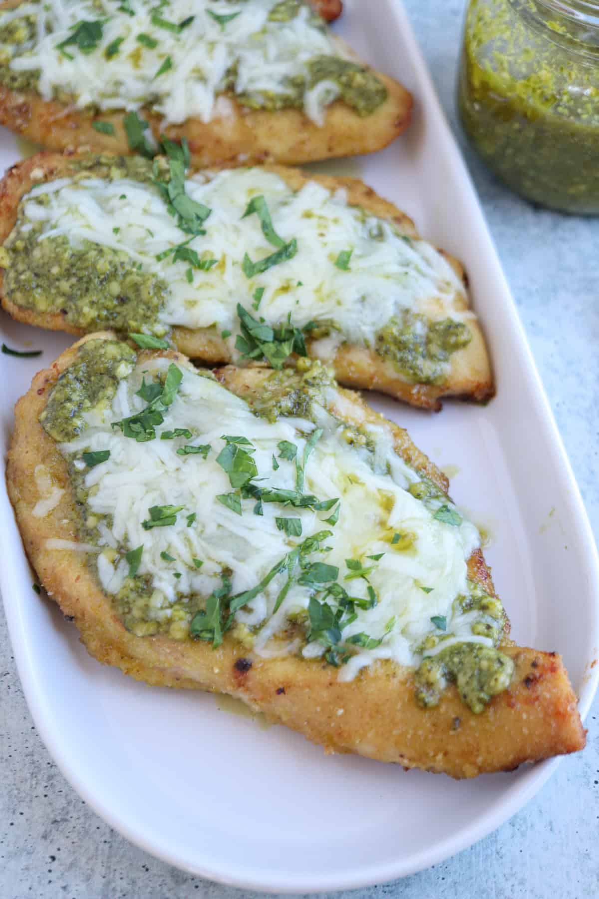 Three pieces of baked pesto chicken parmesan are topped with green pesto sauce, melted cheese, and chopped fresh herbs, arranged on a white rectangular plate with a jar of pesto visible in the background.